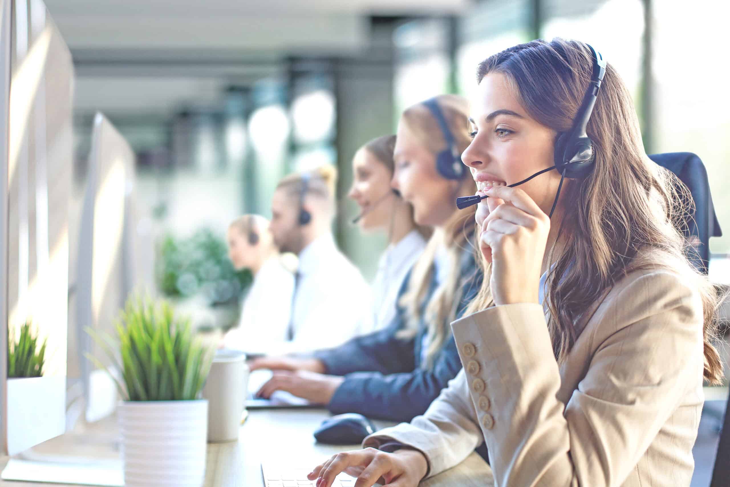 Female customer support operator with headset and smiling, with collegues at background.