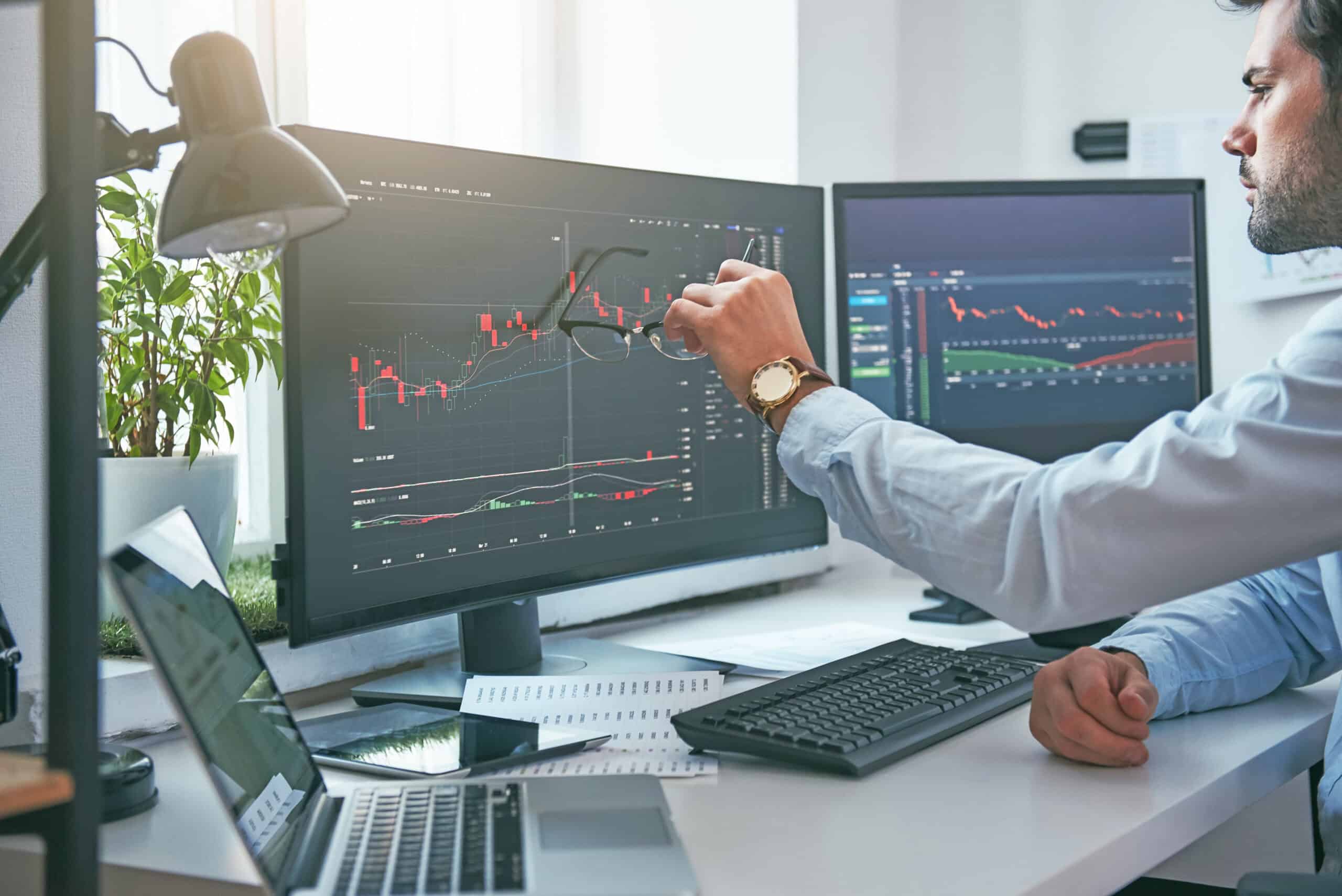 Young exec is pointing at graphs on computer screen and analyzing data while working in his modern office.